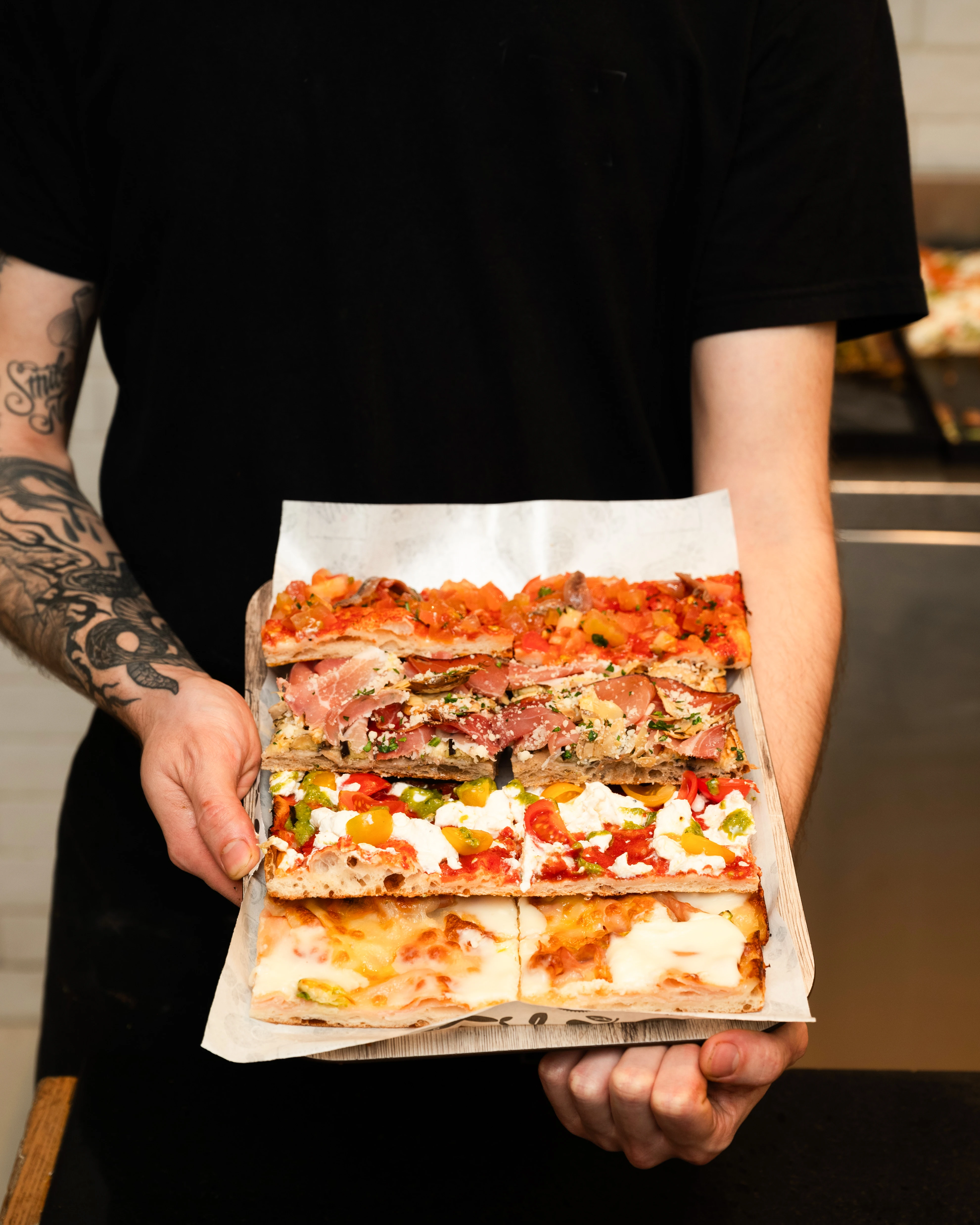 Eine Person in schwarzer Kleidung hält ein Tablett mit frisch gebackenen, verschiedenen Pizza al taglio Stücken.